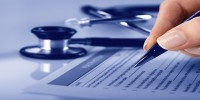 Hand holding a pen, filling in a form with a stethoscope in the background