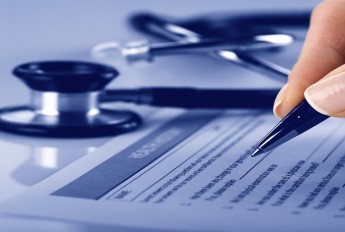 Hand holding a pen, filling in a form with a stethoscope in the background